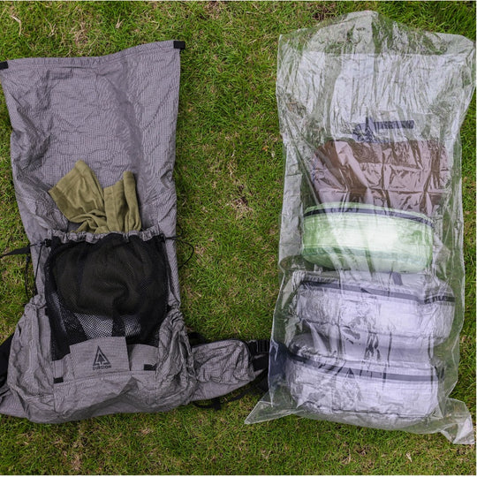 Top-down view of an open backpack next to a clear polycro liner filled with packed gear on grass